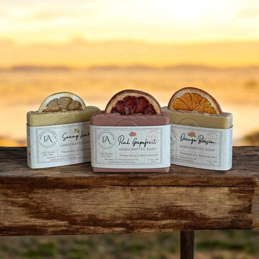 Natural fruit bundle soaps featuring Sunny Lemon, Pink Grapefruit, and Orange Blossom scents on a wooden surface with a sunset backdrop.