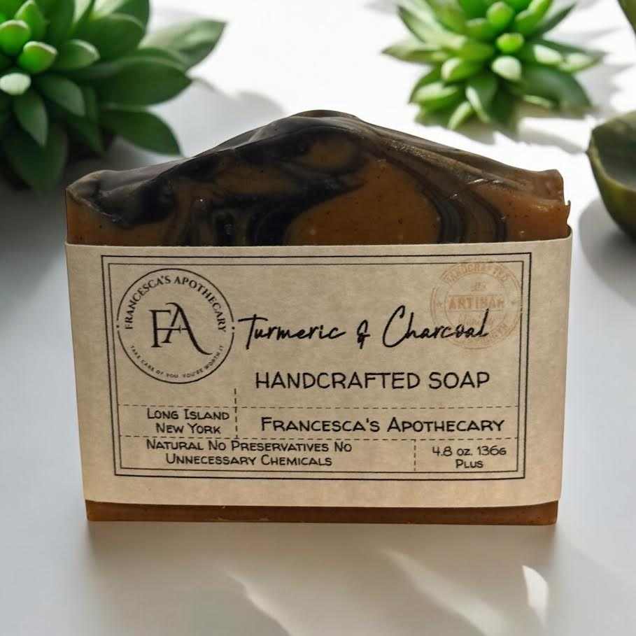 Charcoal & Turmeric Soap bar displayed on a white surface with plants, featuring handcrafted natural ingredients.